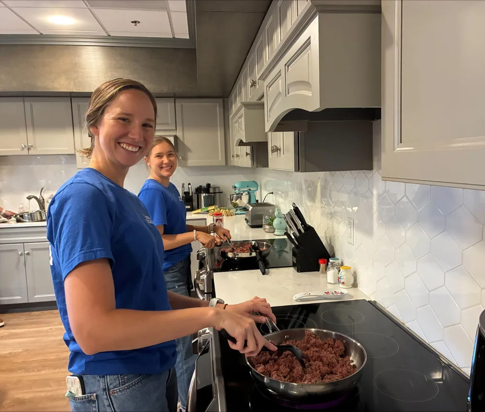 Two volunteers smiling while cooking a meal in the House kitchen.
