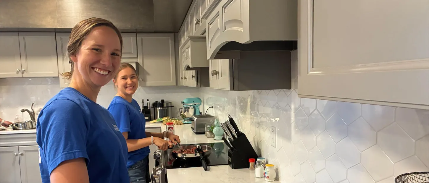 Two volunteers smiling while cooking a meal in the House kitchen.