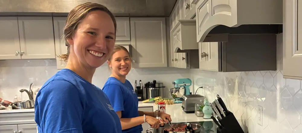 Two volunteers smiling while cooking a meal in the House kitchen.